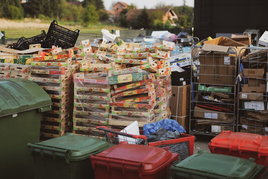 The image displays an outdoor waste collection area with a large stack of flattened cardboard fruit and vegetable packaging boxes, with vibrant red, green, and yellow branding, arranged in multiple vertical stacks. To the left, there are large green wheeled bins with closed lids, situated on a paved surface. In the background, various black plastic crates and metal shelving units hold a variety of boxed items and packages, some labeled with product descriptions and barcodes. On the right side, an orange plastic box and a shopping trolley are visible, partially filled with bags and loose items. The environment appears to be an industrial or commercial waste collection site, with an open space and distant buildings and trees under natural daylight. Waste Removal Waterloo likely manages the collection and disposal of these items, highlighting an example of private bulk waste clearance services for premises such as flats or communal living complexes.