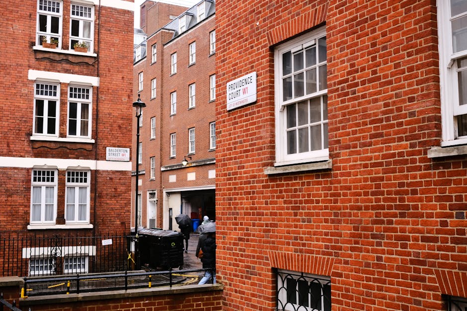 A narrow urban alleyway situated between red brick residential buildings in a densely populated area. The foreground features a brick wall with a white-framed double-hung sash window and a small black iron security grill covering a basement window below. To the left, a black metal fence interrupts the scene, with two waste collection bins positioned near the alley's entrance, one black and the other grey, each with lids. Two individuals in dark clothing are visible, one holding a plastic bag, possibly involved in rubbish disposal. In the background, additional tall brick buildings with multiple white-framed windows extend into the distance. A street sign labeled 'Balderton Street W1' attaches to the brick wall on the left, and another sign reading 'Providence Court W1' is mounted on the building on the right, indicating the area. The scene is lit with natural daylight, creating a clear and neutral atmosphere consistent with on-site waste collection or private rubbish removal activities, as offered by Waste Removal Waterloo.