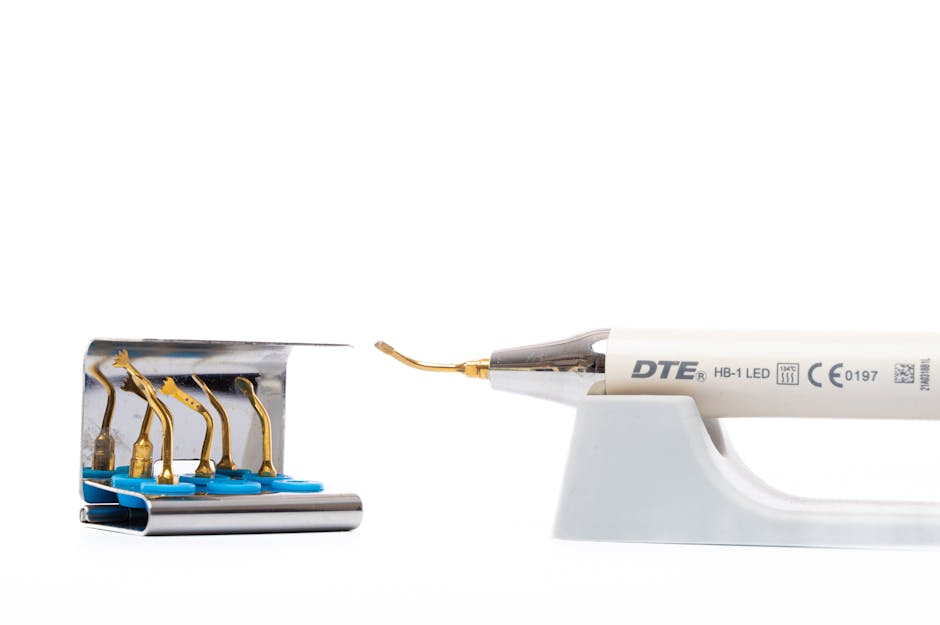 A close-up image of a dental curing light, specifically a DTE HB-1 LED model, positioned on a small white stand. Its slender, white handle features grey text and symbols indicating technical specifications and certifications. The tip of the light is extended forward, displaying a small, curved optical lens made of transparent material. To the left of the curing light is a metallic tray or holder with multiple small, gold-colored dental curing light tips, each attached to a short, curved metallic arm, with their bases secured to a shiny, reflective metal surface. The background is plain white, highlighting the precision and cleanliness associated with dental tools. This setup appears to be part of a dental procedure or dental equipment display, aligning with the context of professional clinical environments, commonly requiring maintenance or disposal of such small medical devices, similar to those handled by waste removal services like Waste Removal Waterloo in the UK.