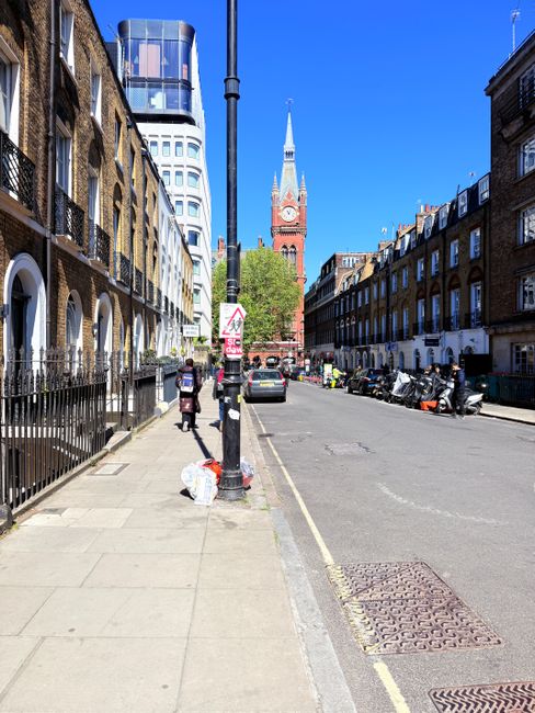 A city street in London during daytime, featuring a row of traditional brick terraced houses with white window frames and black iron railings lining the left side, and a wide sidewalk with a metal pole supporting a traffic sign and a standing pedestrian. On the sidewalk, there are two people walking past a plastic bag of rubbish and a small pile of waste beside the pole. The street is also occupied by parked motorcycles and small cars along the curb on the right, with some construction or maintenance workers visible further down. In the background, the iconic clock tower of the Houses of Parliament, made of red brick with a tall spire, rises beneath a bright blue sky, complemented by modern office buildings with reflective glass facades and a distinctive rounded corner structure. The scene illustrates a typical urban environment where private waste collection or rubbish removal services, such as Waste Removal Waterloo, might operate to manage local refuse and ensure clean, tidy streets in central London.