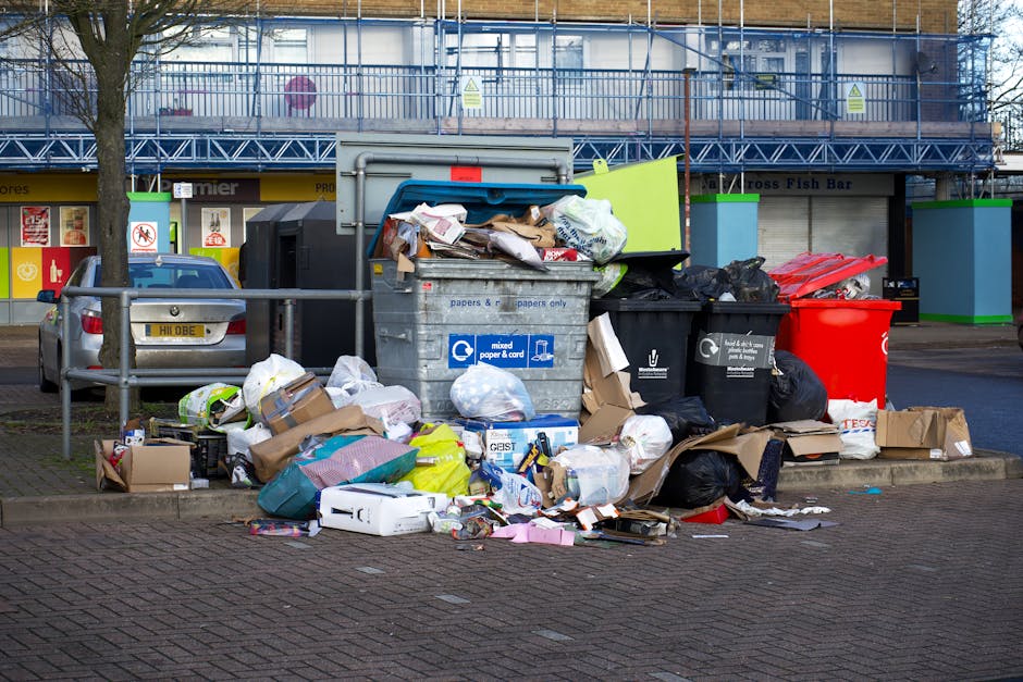 A collection of overflowing rubbish bins and scattered waste including cardboard boxes, plastic bags, paper, and packaging materials positioned on a paved area adjacent to a parking lot. The scene features a large grey mixed waste bin with a partly open blue lid, placed in front of a metal railing and nearby a smaller black bin and a prominent red bin. Several paper and plastic items, some crumpled or flattened, spill onto the pavement in front of the bins. Behind the waste area, there is a background of commercial premises with closed shop shutters, signage, and a building with scaffolding and blue safety netting, suggesting ongoing construction or maintenance. The setting appears to be outdoors during daytime, with natural lighting illuminating the scene, reflecting a typical urban environment where private waste collection by services such as Waste Removal Waterloo may be necessary to manage rubbish outside standard council collections.