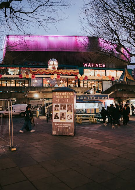 The image depicts an outdoor scene in front of a busy market with several people walking and standing on a paved area. Prominently in the foreground, there is a large, freestanding wooden information or advertising board displaying various images and text related to local events or announcements. To the right, a small food stall or kiosk labeled 'Mexican Street Kitchen' with a blue and white striped canopy is visible, with a few customers gathered nearby. In the background, a multi-level building features large glass windows and a colourful, decorative exterior, including a series of orange and pink floral banners and a bright, illuminated sign spelling 'WAHACA.' The building's upper section is covered by a distinctive overhanging roof with a pink and purple hue, illuminated from behind. The scene is set during dusk or early evening, with soft, ambient lighting and leafless tree branches framing the upper edges of the image, suggesting a winter or early spring setting. The overall environment appears lively and urban, consistent with a marketplace or entertainment venue, with no visible waste or rubbish, implying it is a well-maintained area managed by services such as Waste Removal Waterloo that handle private waste collection and on-site clearance for commercial and public spaces.