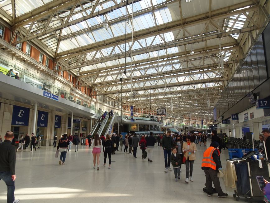 Inside a spacious train station with a high, arched glass ceiling supported by metal trusses allowing natural light to illuminate the area. The wide, polished floor is populated with numerous travelers walking in various directions, some with luggage trolleys or carrying bags. In the foreground, a station staff member wearing a high-visibility orange vest is seen handling a collection of discarded items or cleaning equipment near a row of lockers or a luggage area. On the left side, a group of people wait in front of ticket counters and entrance signs, while on the right, a large digital departure board displays information. An escalator ascends toward an upper level with more travelers visible. The environment appears busy yet orderly, typical of a major transportation hub, with a focus on passenger movement and station operations, reflecting a context where private rubbish or waste removal services may be relevant for station cleaning or maintenance work. The setting emphasizes the functional, public environment of a busy railway station often serviced by waste removal professionals for maintaining cleanliness and hygiene.