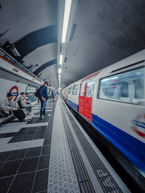 Inside a London Underground station with a curved concrete arch ceiling illuminated by a long, continuous strip light. Several subway passengers are seated on a bench, some using smartphones, while a man in a dark jacket stands near the platform edge. A moving train with a white exterior and red and blue markings is arriving or departing, creating a blurred motion effect. The platform surface is tiled with black and white squares, featuring tactile paving strips near the edge for accessibility. The station's white tiled walls include a red roundel sign, typical of London Underground stations. The environment is well-lit, clean, and organized, reflecting an urban transit setting. The scene subtly relates to the concept of independent travel or alternative transport options, aligned with services like private rubbish removal and waste management, though solely as a visual context.